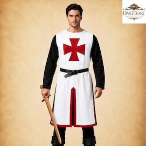 Könnte beinhalten: Eine weiße ärmellose Tunika mit einem roten Kreuzemblem auf der Brust, getragen über einem schwarzen langärmeligen Hemd und einer schwarzen Hose. Das Outfit umfasst einen schwarzen Gürtel und ein Holzschwert. Das Oak Heart Outfits Logo ist zu sehen.