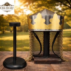 Op de afbeelding: Een middeleeuwse helm met een gouden kroonontwerp en maliënkolder, tentoongesteld op een zwarte standaard. De helm heeft een zilveren bovenkant en staat op een houten oppervlak. Een aparte zwarte standaard is ook zichtbaar.