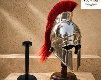 May include: A polished silver-toned metal helmet with a prominent red crest, mounted on a dark wooden stand. Beside it stands a separate black display stand with a circular base and a vertical post. The text Oak Heart Outfits is visible.