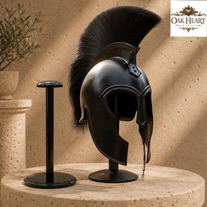 May include: A matte black Spartan-style helmet featuring a prominent black crest made of long, stiff bristles. The helmet is mounted on a black metal display stand, with a matching empty black stand positioned directly to its left.