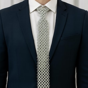 May include: A silver chainmail tie worn over a white dress shirt and a navy blue suit jacket. The tie is made of small, interlocking metal rings, creating a unique, textured appearance. The suit jacket has a classic two-button design.