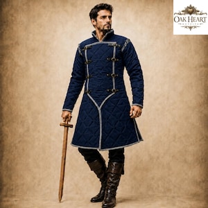 May include: A dark blue quilted gambeson featuring a high collar, grey trim, and four horizontal buckle straps across the chest. The garment has long sleeves and a knee-length hem. A wooden sword is held in one hand. The logo Oak Heart Outfits is visible.