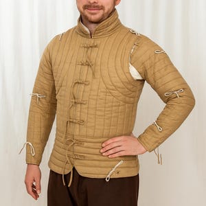 May include: A tan quilted gambeson, a historical garment with a high collar and long sleeves. It features vertical stitching, tie fastenings, and white accents. This protective layer is designed for medieval reenactment or costume.