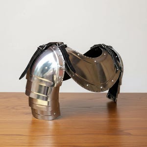 Pode incluir: Uma pe&ccedil;a de armadura de ombro em a&ccedil;o polido com placas em camadas e um design curvo. A armadura &eacute; fixada com correias de couro preto e rebites, refletindo a luz. Esta armadura de estilo medieval foi projetada para proteger o ombro.