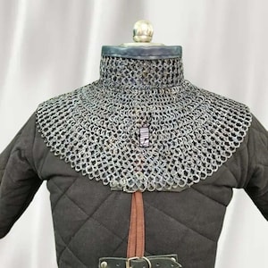 May include: A medieval chainmail collar, crafted from interlocking metal rings, is displayed over a black quilted gambeson. The chainmail protects the neck and shoulders. A leather strap and buckle are visible at the base of the gambeson.