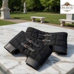 May include: Black quilted leg armor with leather accents and brass buckles. The armor is designed with a diamond pattern and is displayed on a marble surface. The Oak Heart Outfits logo is in the top right corner.