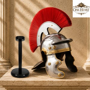 May include: A silver Roman-style helmet with a prominent red crest and gold-toned trim, featuring decorative circular accents. Beside it stands a black cylindrical display stand with a circular base. The text OAK HEART OUTFITS is visible.