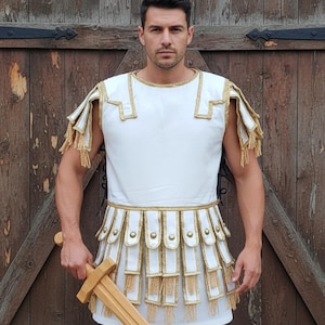 May include: A white Roman-style tunic with gold trim and fringe. The costume includes shoulder pieces and a skirt-like lower section. A wooden sword is held in the hand. The outfit is designed to resemble ancient Roman attire.