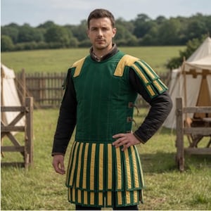 May include: Green and yellow medieval gambeson with vertical stripes. The padded garment features yellow shoulder accents and a matching skirt-like bottom. The person is wearing a black long-sleeved shirt underneath. The background shows a field and tents.