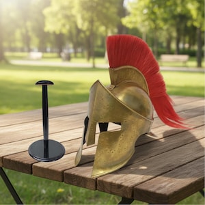 Könnte beinhalten: Ein goldfarbener spartanischer Helm mit roter Feder sitzt auf einem Holztisch. Ein schwarzer Helmhalter befindet sich links. Der Hintergrund ist eine verschwommene Außenszene mit Bäumen und Gras.