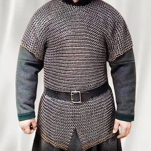 May include: A chainmail shirt with a black leather belt and buckle. The shirt has long grey sleeves and a black collar. The chainmail is made of small, interlocking metal rings. The shirt is worn over a grey tunic.