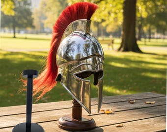 May include: A polished silver Spartan helmet with a vibrant red plume, displayed on a wooden stand. The helmet features cheek guards and a detailed design. A separate black stand is also visible, set against a blurred outdoor background.