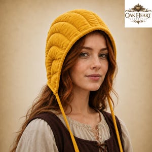 May include: A mustard yellow quilted bonnet featuring a rounded, multi-paneled design with long fabric ties hanging down. The headwear has a textured, soft appearance and frames the face, with the Oak Heart Outfits logo visible in the corner.