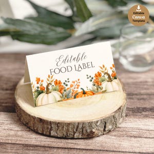May include: A folded card with the text "Editable FOOD LABEL" and a watercolor design of pumpkins and fall foliage. The card is displayed on a wooden slice, with a blurred background of greenery and a glass of water.