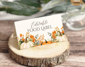 May include: A folded card with the text "Editable FOOD LABEL" and a watercolor design of pumpkins and fall foliage. The card is displayed on a wooden slice, with a blurred background of greenery and a glass of water.