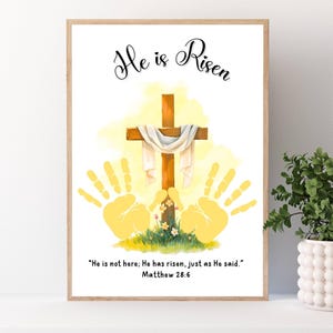 May include: A framed print with the words "He is Risen" in elegant script. The artwork features a wooden cross draped with a white cloth, yellow handprints, and the Bible verse Matthew 28:6. The background has a soft yellow glow.