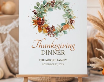 May include: A white sign on a wooden easel with the words "Welcome To Our Thanksgiving Dinner" and the family name and date. The sign is decorated with a wreath of leaves and berries. Pumpkins and decorative plants are in the background.