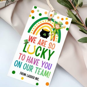 Może przedstawiać: Etykieta prezentowa na Dzień Świętego Patryka z tęczą, skrzatem i ilustracją garnka ze złotem. Etykieta głosi: „Jesteśmy szczęśliwi, że jesteś w naszym zespole!” i zawiera tekst „Od: Ladder Inc.”