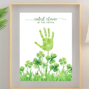 Peut inclure: Impression d'art encadrée avec une empreinte de main verte et des trèfles. Le texte "cutest clover in the patch" est écrit au-dessus de l'empreinte. L'œuvre présente une empreinte de main verte au-dessus d'un champ de trèfles et d'herbe verts. Le cadre est marron clair.