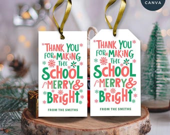 Könnte beinhalten: Zwei weiße Geschenkanhänger mit goldenem Band, mit dem Text "Thank You for Making the School Merry & Bright" in Rot und Grün. Die Anhänger liegen auf einer Holzscheibe, mit verschwommenem Feiertagshintergrund.
