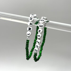 K&ouml;nnte beinhalten: Zwei gr&uuml;ne Perlenarmb&auml;nder mit wei&szlig;en Buchstabenperlen, die "CUZ SHE'S" und "LWYMM" buchstabieren.