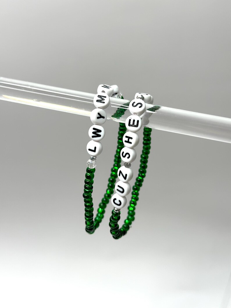 K&ouml;nnte beinhalten: Zwei gr&uuml;ne Perlenarmb&auml;nder mit wei&szlig;en Buchstabenperlen, die "CUZ SHE'S" und "LWYMM" buchstabieren.