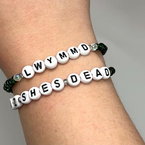 K&ouml;nnte beinhalten: Zwei Perlenarmb&auml;nder mit wei&szlig;en Perlen und schwarzen Buchstaben. Das obere Armband buchstabiert "LWYMMD" und das untere Armband buchstabiert "ZSHESDEAD". Beide Armb&auml;nder haben gr&uuml;ne und klare Perlen.