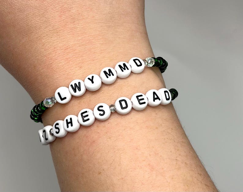 K&ouml;nnte beinhalten: Zwei Perlenarmb&auml;nder mit wei&szlig;en Perlen und schwarzen Buchstaben. Das obere Armband buchstabiert "LWYMMD" und das untere Armband buchstabiert "ZSHESDEAD". Beide Armb&auml;nder haben gr&uuml;ne und klare Perlen.