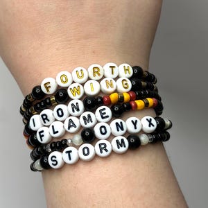 May include: A stack of seven beaded bracelets with black and white beads. The beads spell out the words "Fourth Wing", "Iron Flame", "Onyx", and "Storm".