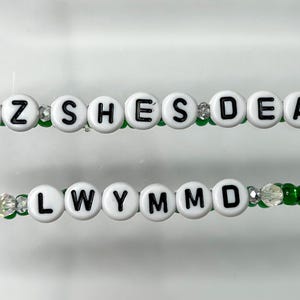 K&ouml;nnte beinhalten: Zwei Perlenarmb&auml;nder mit wei&szlig;en Buchstabenperlen, die "CUZ SHE'S DEAD" und "LWYMMD" auf gr&uuml;ner Schnur buchstabieren.