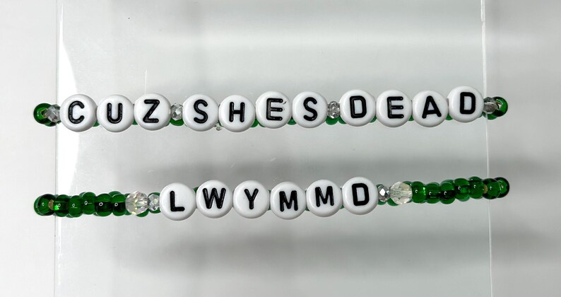K&ouml;nnte beinhalten: Zwei Perlenarmb&auml;nder mit wei&szlig;en Buchstabenperlen, die "CUZ SHE'S DEAD" und "LWYMMD" auf gr&uuml;ner Schnur buchstabieren.