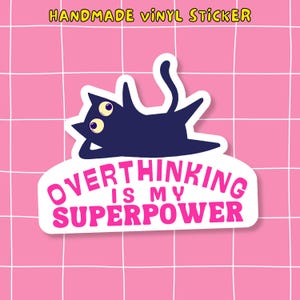 May include: A handmade vinyl sticker with a navy blue cat illustration, three yellow eyes, and the text "OVERTHINKING IS MY SUPERPOWER" in pink. The sticker is on a white background, set against a pink grid pattern.