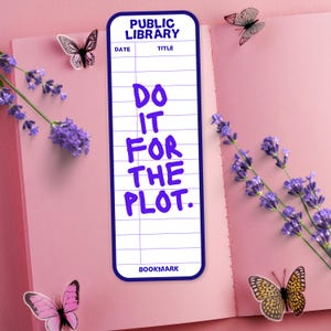 May include: A bookmark with a blue border and the text "DO IT FOR THE PLOT" in purple. The bookmark is designed to look like a library card. The image also features lavender sprigs and butterfly stickers on a pink book.