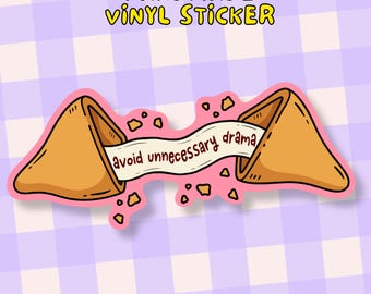 May include: A handmade vinyl sticker featuring a cartoon fortune cookie with the message "avoid unnecessary drama" on a banner. The sticker has a pink outline and is set against a checkered background.