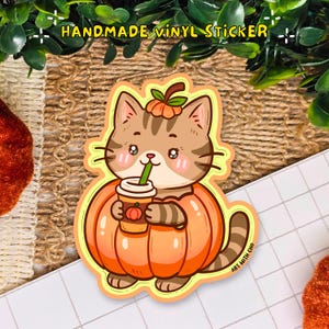 May include: A handmade vinyl sticker showing a cartoon cat in a pumpkin costume, holding a coffee cup with a green straw. The cat is brown and tan with pink cheeks. The pumpkin is orange with a green stem.