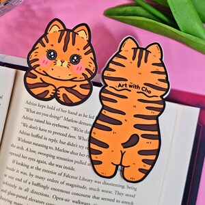 May include: Two orange and brown tiger-striped cat-shaped bookmarks. One shows the cat's face with a cute expression, and the other shows the cat's back. The text "Art with Cho" is on the back of the bookmark. The bookmarks are on top of an open book.
