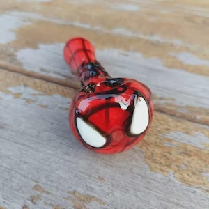 May include: A red and black glass pipe featuring a web-like pattern across the surface and two prominent white, almond-shaped eye designs on the front. The object has a smooth, glossy finish and a rounded bowl shape with a tapered stem.