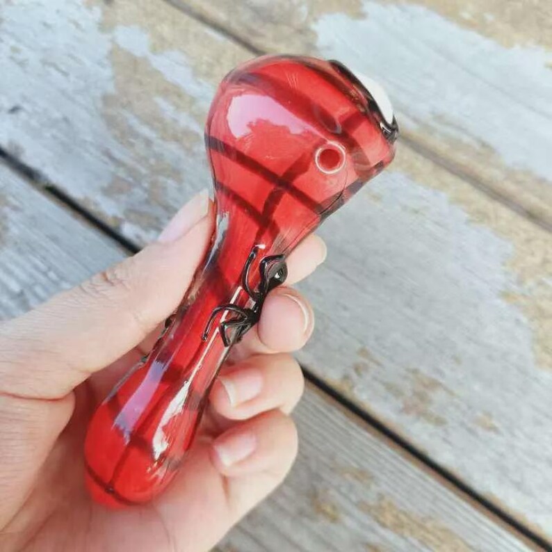 May include: A red glass pipe featuring a translucent finish with darker red stripes and a small black spider figurine attached to the side of the stem. The pipe has a rounded bowl at the top and a circular carb hole on the side of the bowl area.