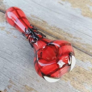 May include: A red glass pipe featuring a black spider decoration on the stem and two white accents on the bowl. The pipe has a smooth, glossy finish with a translucent red body and dark red swirling patterns throughout the glass structure.
