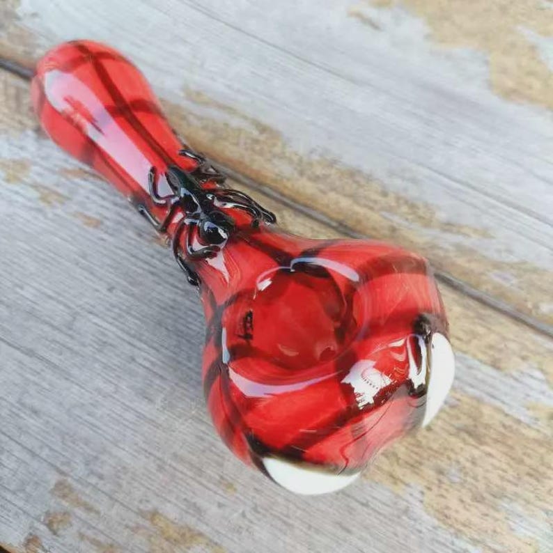 May include: A red glass pipe featuring a black spider decoration on the stem and two white accents on the bowl. The pipe has a smooth, glossy finish with a translucent red body and dark red swirling patterns throughout the glass structure.