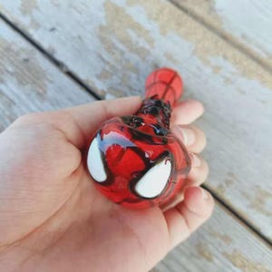 May include: A red and black glass pipe featuring a rounded bowl with two large white eye-shaped accents. The surface of the glass is decorated with a dark web-like pattern that extends from the bowl along the cylindrical stem of the object.