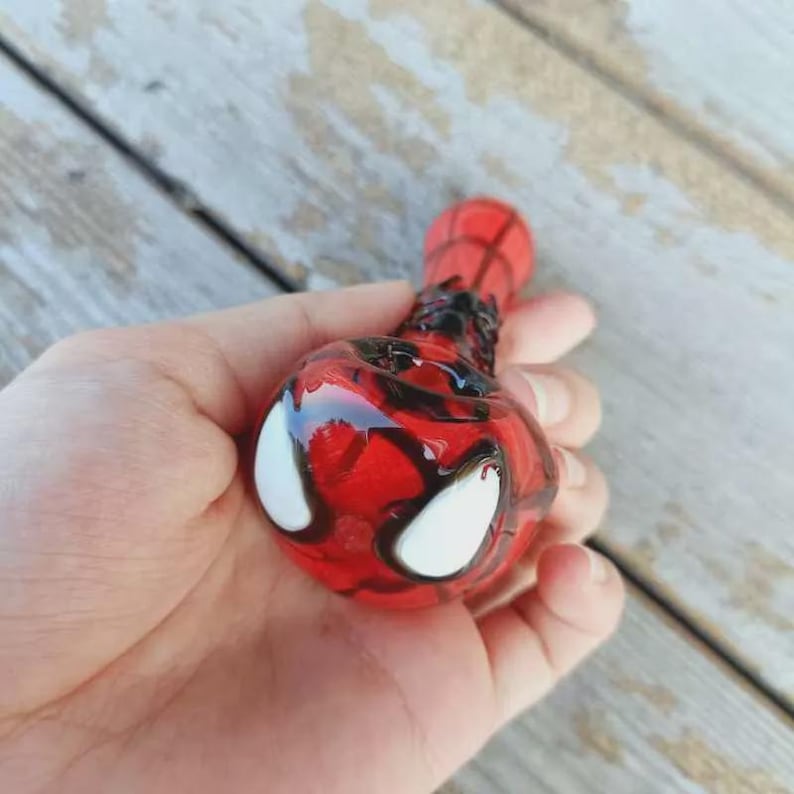 May include: A red and black glass pipe featuring a rounded bowl with two large white eye-shaped accents. The surface of the glass is decorated with a dark web-like pattern that extends from the bowl along the cylindrical stem of the object.