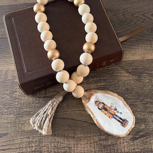 May include: A wooden bead garland with gold accents and a tassel hangs from a painted oyster shell with a nutcracker design. The garland is draped over a brown leather-bound book.