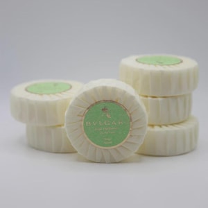 May include: Several round, white soaps with a pleated paper wrapping are stacked and arranged on a white background. Each soap has a green label with gold text. The label reads "BVLGARI" and "Eau Parfumée au Thé Vert".