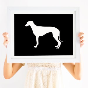 May include: A white silhouette of a dog, possibly a Greyhound, is printed on a black background. The print is framed in a white frame and is being held by a person.