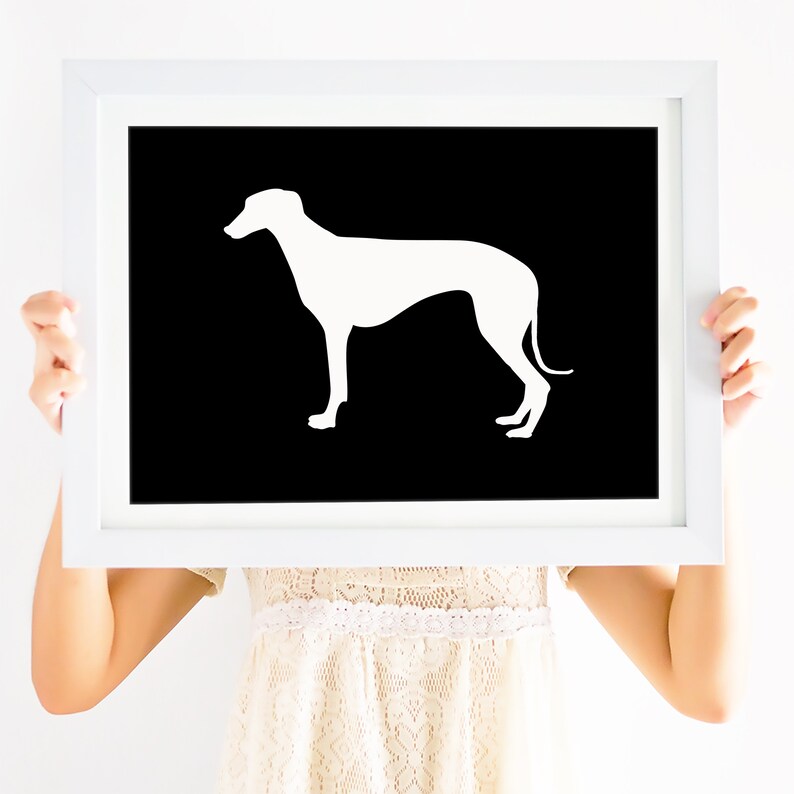 May include: A white silhouette of a dog, possibly a Greyhound, is printed on a black background. The print is framed in a white frame and is being held by a person.