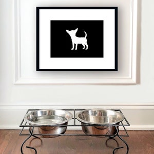 May include: A black framed picture with a white silhouette of a chihuahua on a black background. Two silver metal dog bowls are on a black metal stand.