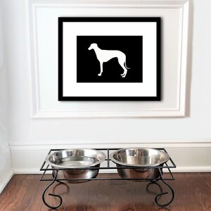 May include: Black and white framed print of a greyhound silhouette on a white background. The print is hanging on a white wall above a black metal double dog bowl stand.
