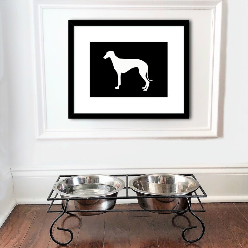 May include: Black and white framed print of a greyhound silhouette on a white background. The print is hanging on a white wall above a black metal double dog bowl stand.