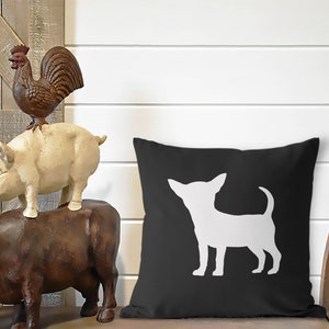 May include: A black throw pillow with a white silhouette of a chihuahua dog. The pillow is sitting on a wooden chair with a brown cow, pig, and rooster figurine stacked on top of each other.